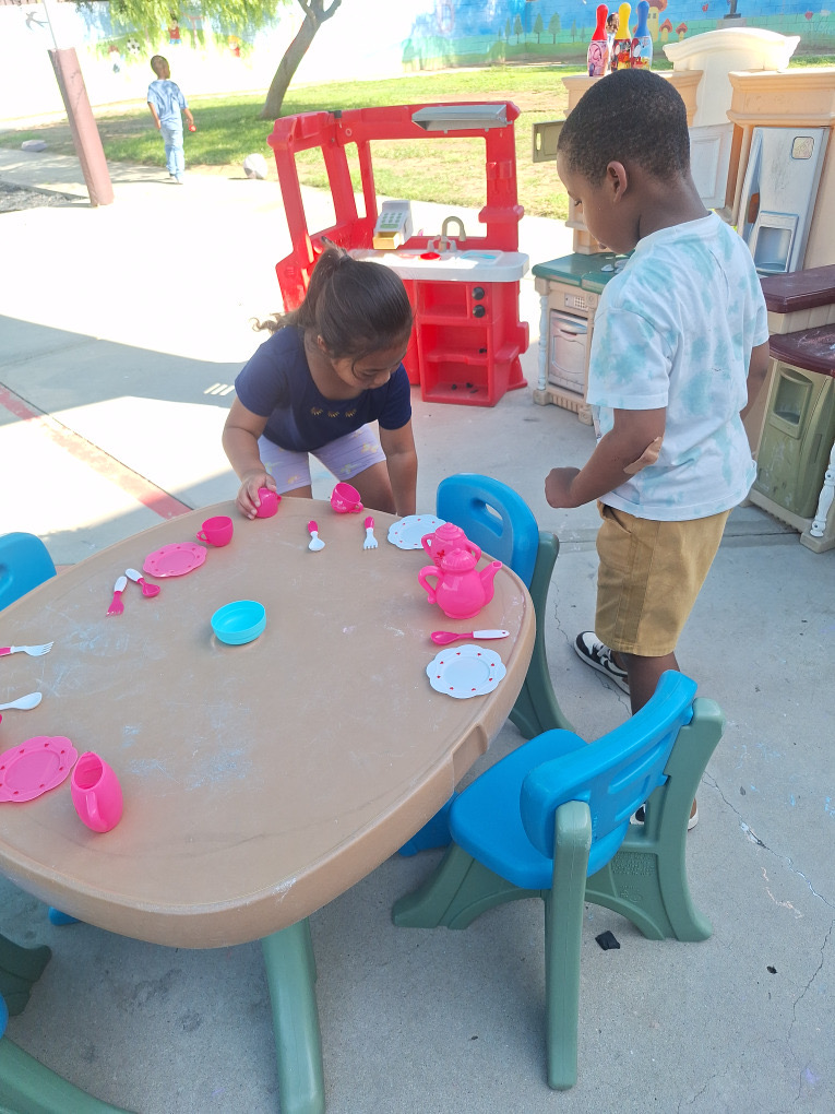Two children arranging toy cups and dishes at an outdoor table